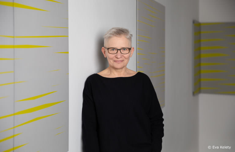 Halbportrait Barbara Höller an Wand lehenend mit Bildern mit gelben schmalen Rautenformen